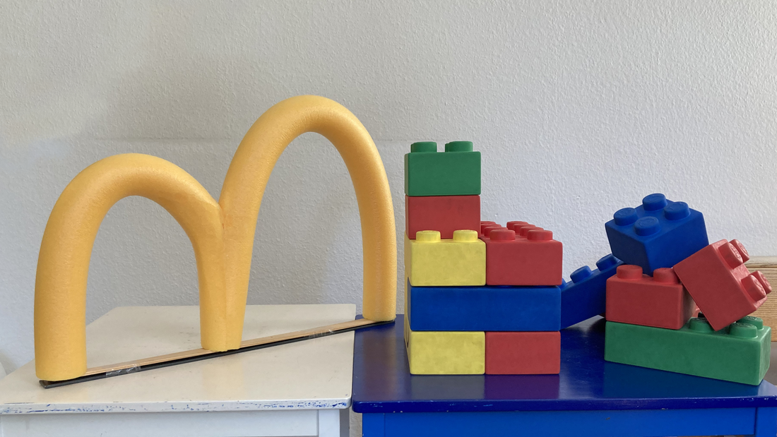 Gelbes "m" steht auf einem Tisch neben großen bunten Legosteinen